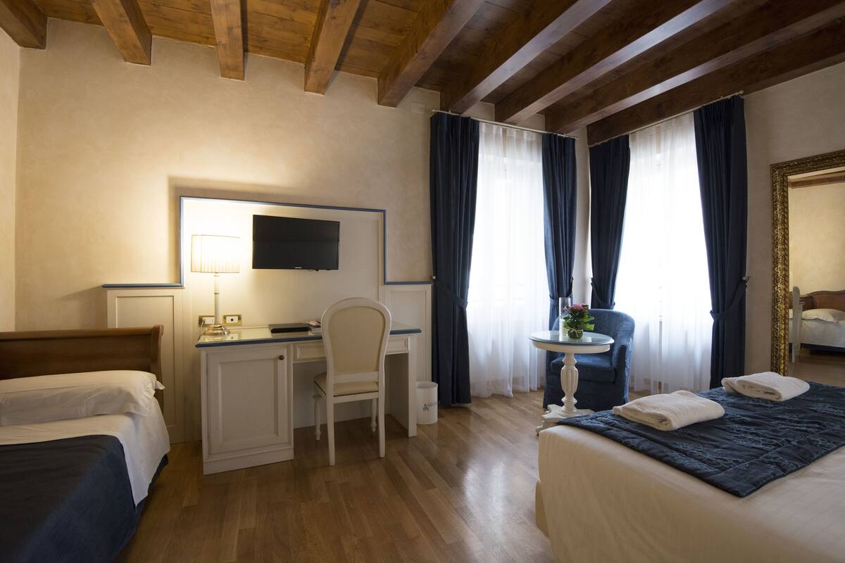 A hotel room with a bed, a desk with a chair, a TV, and a window with curtains.