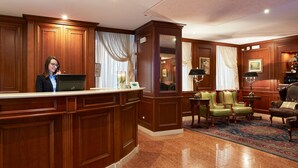 Un área de recepción con un mostrador de madera, una computadora portátil, un sillón verde, un sillón de cuero y una mesita con una lámpara.