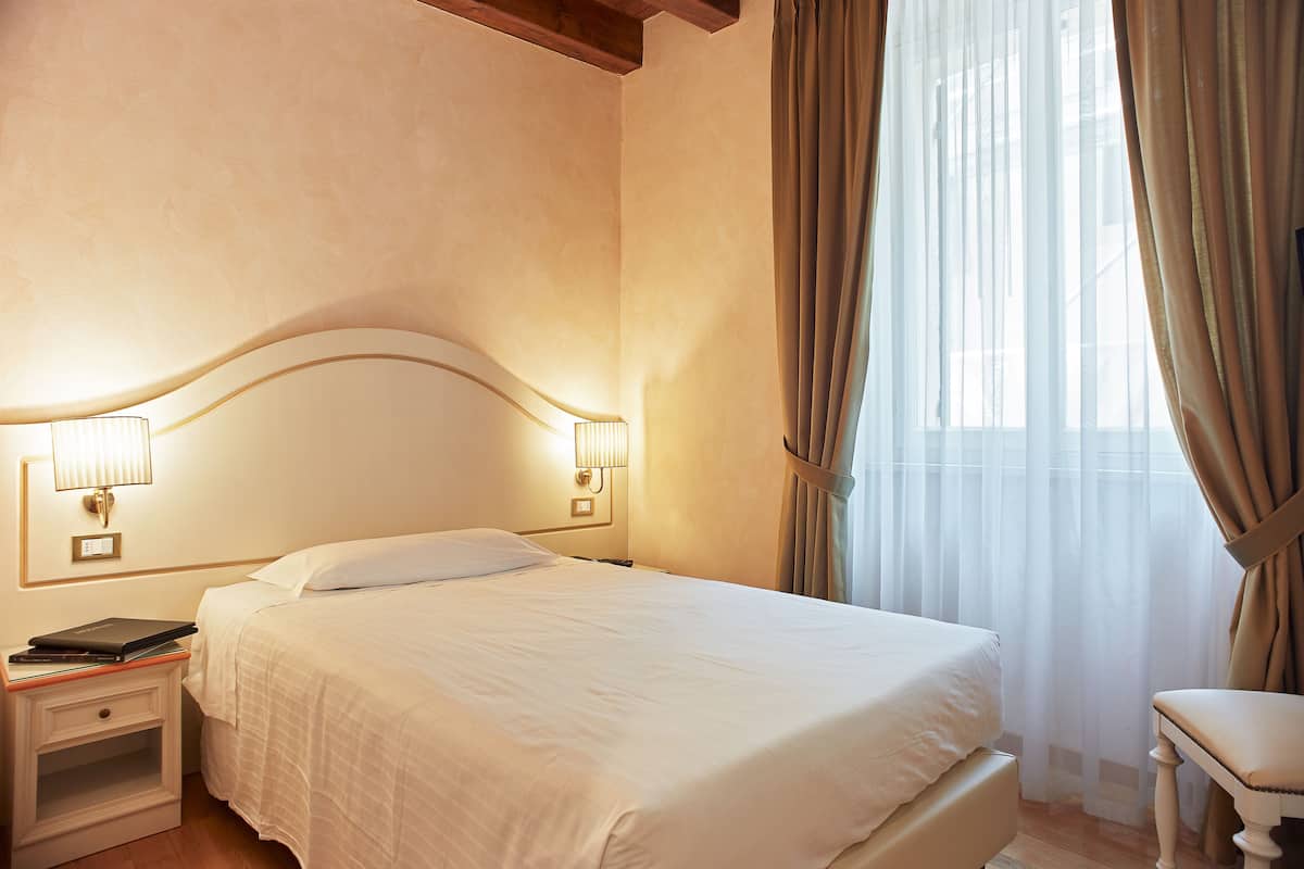 A neatly made bed with a white comforter and pillows, a bedside table with a lamp, and a chair to the right.