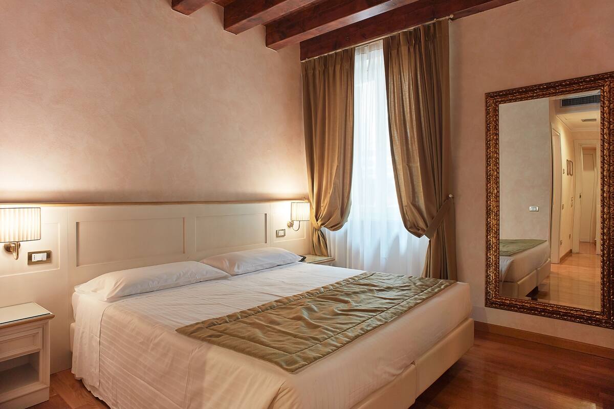 A neatly made bed with a white comforter and a brown throw blanket, a bedside table with a lamp, and a large mirror on the wall.