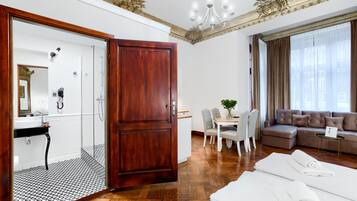 Ein Hotelzimmer mit Holztür, einem Badezimmer inklusive Spiegel, einer Essecke mit Tisch und Stühlen, einem Sofa und einem großen Fenster mit Vorhängen.