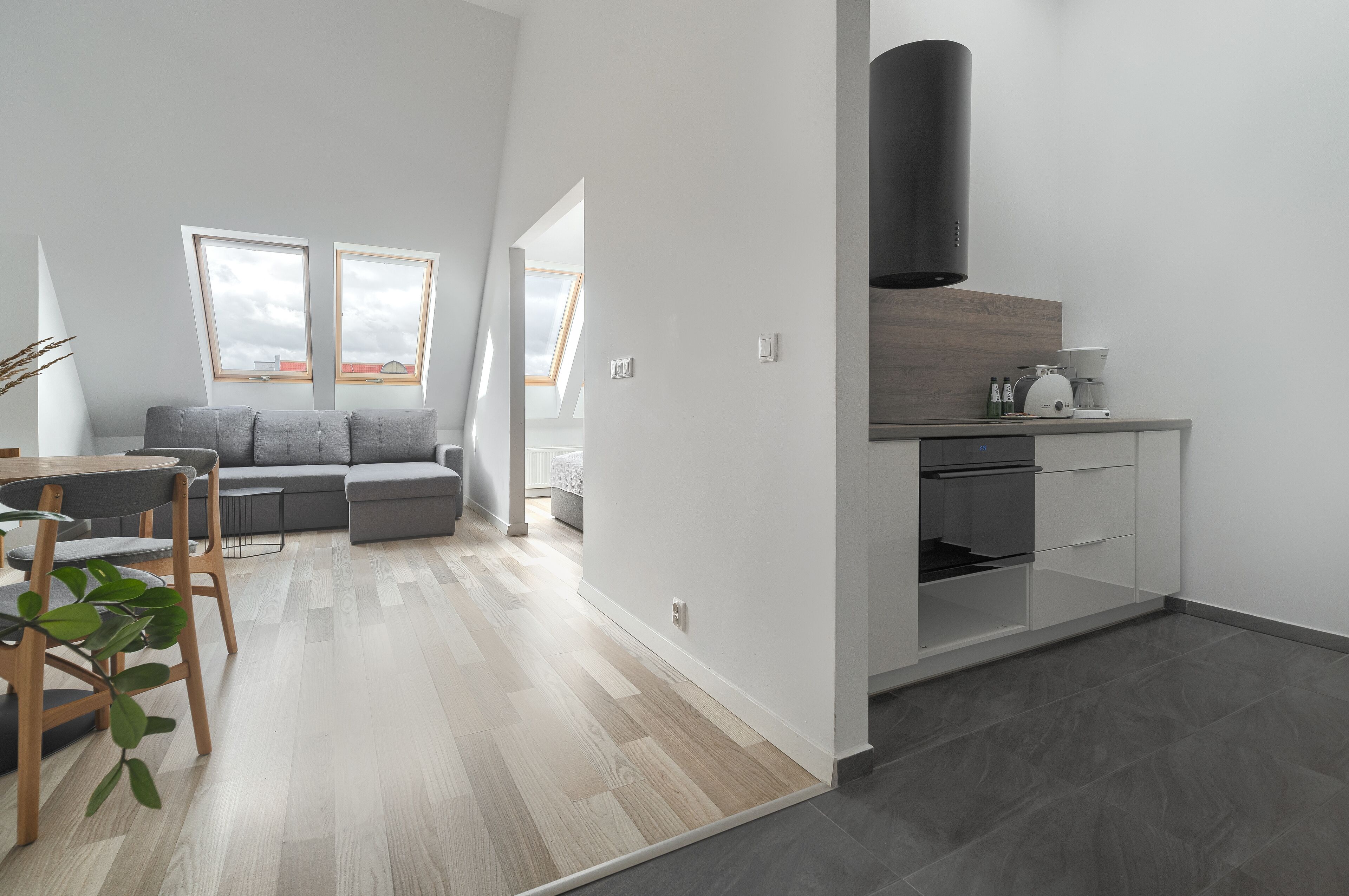 A modern kitchen with a built-in oven and a coffee machine, a dining area with a table and chairs, and a gray sofa in a living area with large windows.