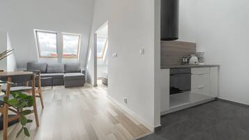 A modern kitchen with a built-in oven and a coffee machine, a dining area with a table and chairs, and a gray sofa in a living area with large windows.