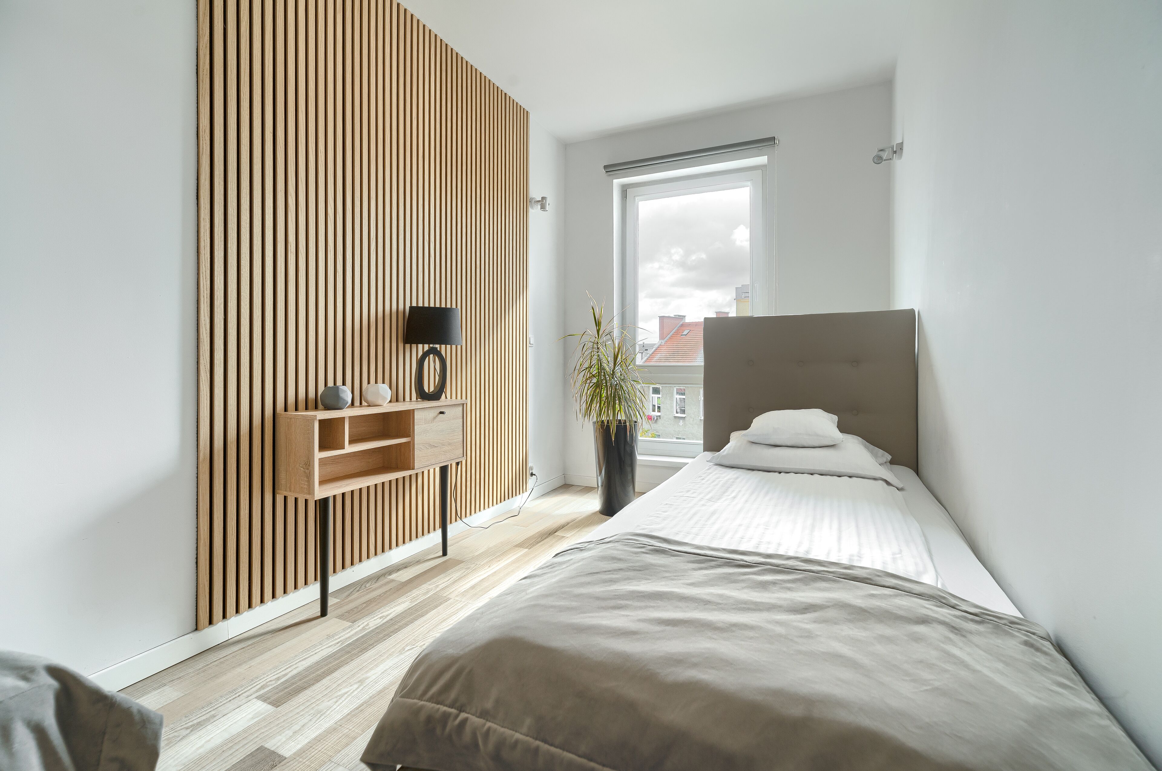 A modern bedroom with a slatted wooden wall, a bedside table with a lamp, a large window, and a bed with white bedding.