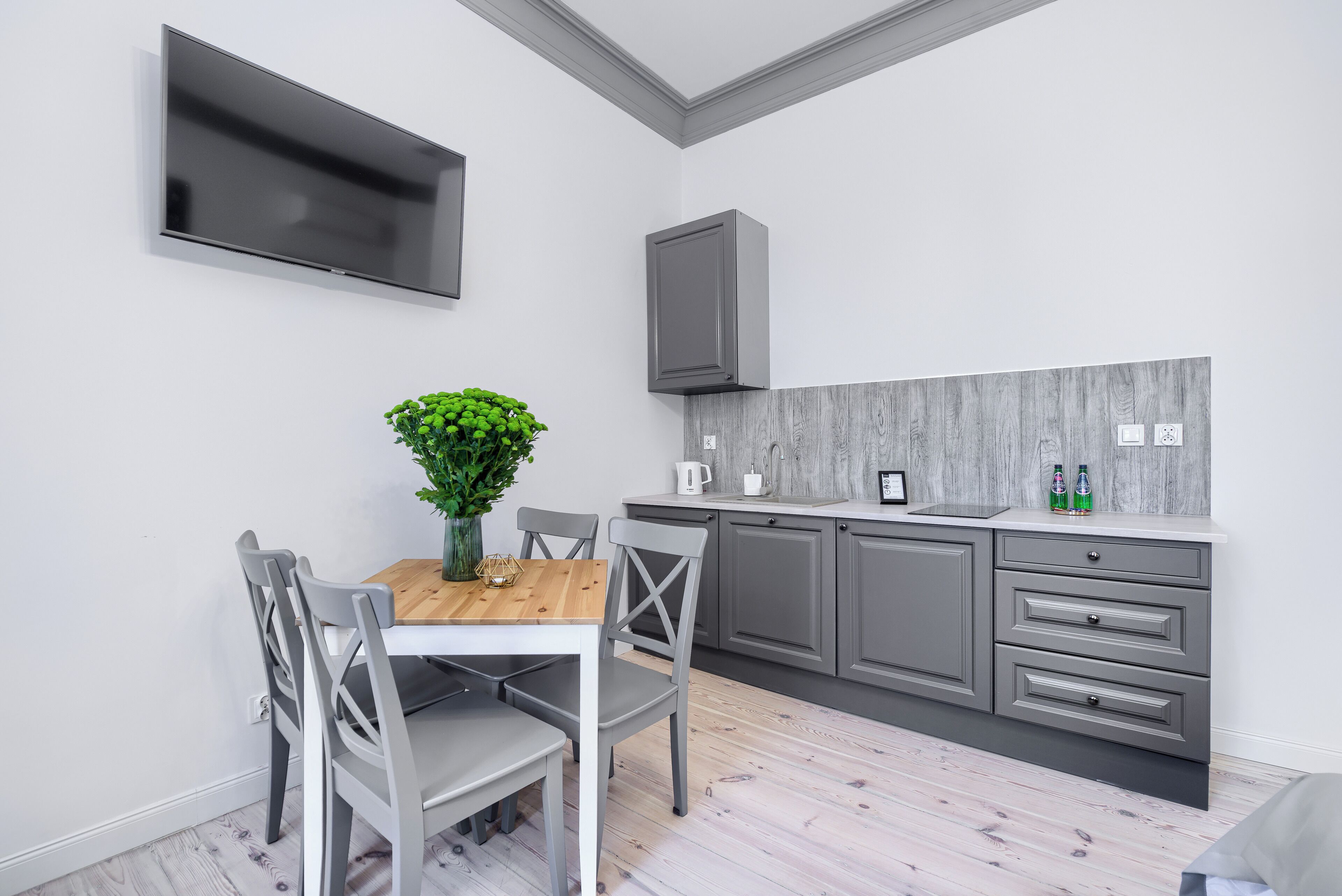 A modern kitchen with grey cabinets, a wooden dining table, and a flat-screen TV mounted on the wall.