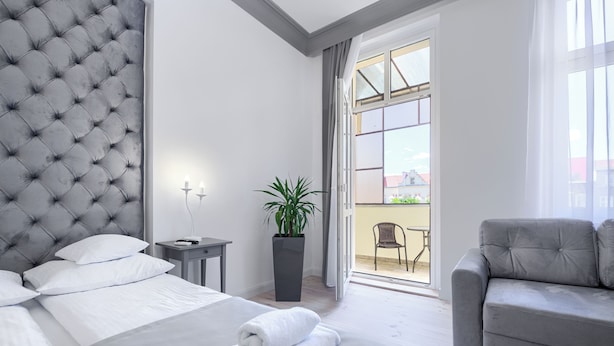 Habitación de hotel con cabecera capitonada, cama con ropa blanca, planta en maceta, sofá gris y balcón con vistas.