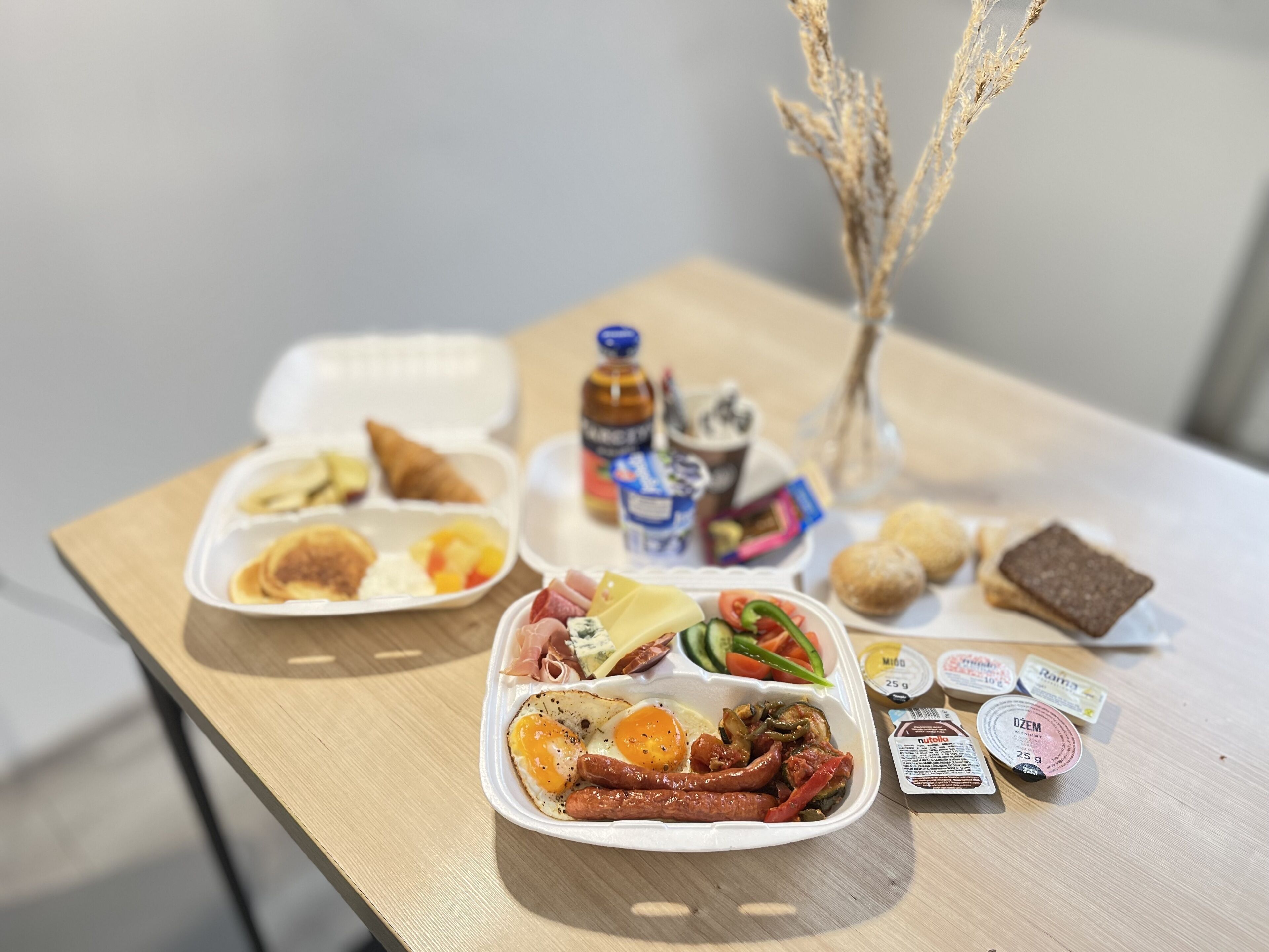 Una mesa con diversos alimentos, incluyendo un sándwich, ensalada y un recipiente con huevos y salchichas. También hay condimentos y una botella de jugo.