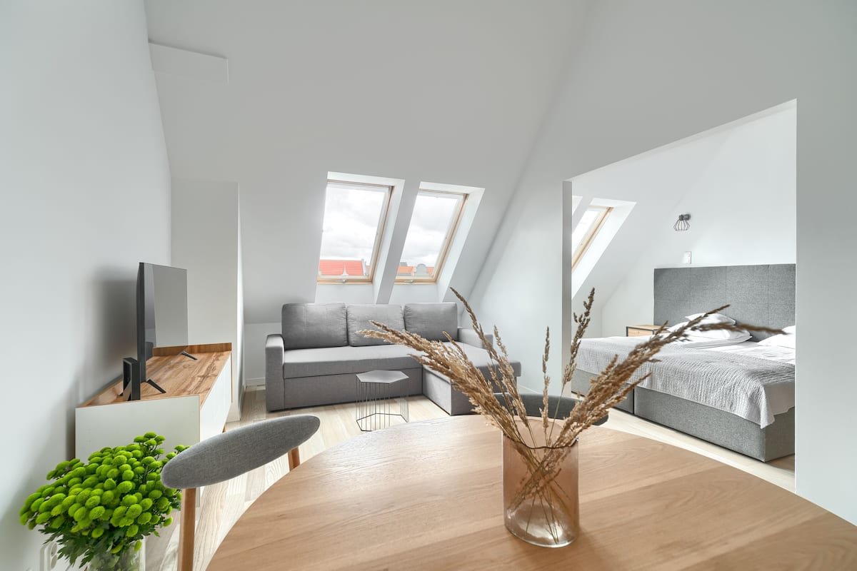 Una habitación moderna y luminosa con una mesa de comedor de madera, un sofá gris, una cama gris y un gran ventanal que proporciona luz natural.