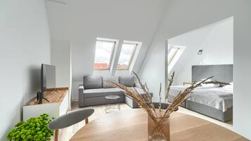 Una habitación moderna y luminosa con una mesa de comedor de madera, un sofá gris, una cama gris y un gran ventanal que proporciona luz natural.
