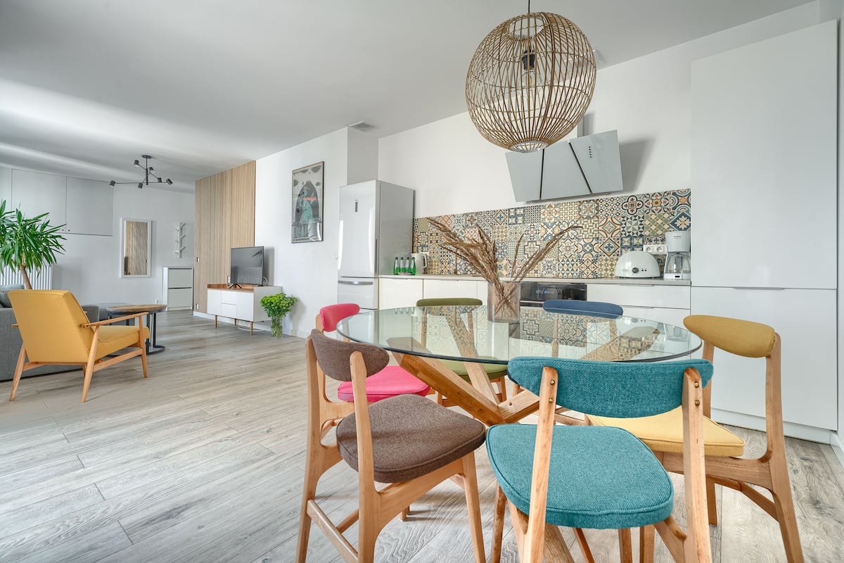 Un comedor moderno con mesa de vidrio y sillas de madera, una cocina con salpicadero decorado y una lámpara colgante.