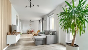 Una sala de estar moderna con un sofá seccional gris, piso de madera, una planta grande en maceta y un comedor al fondo.