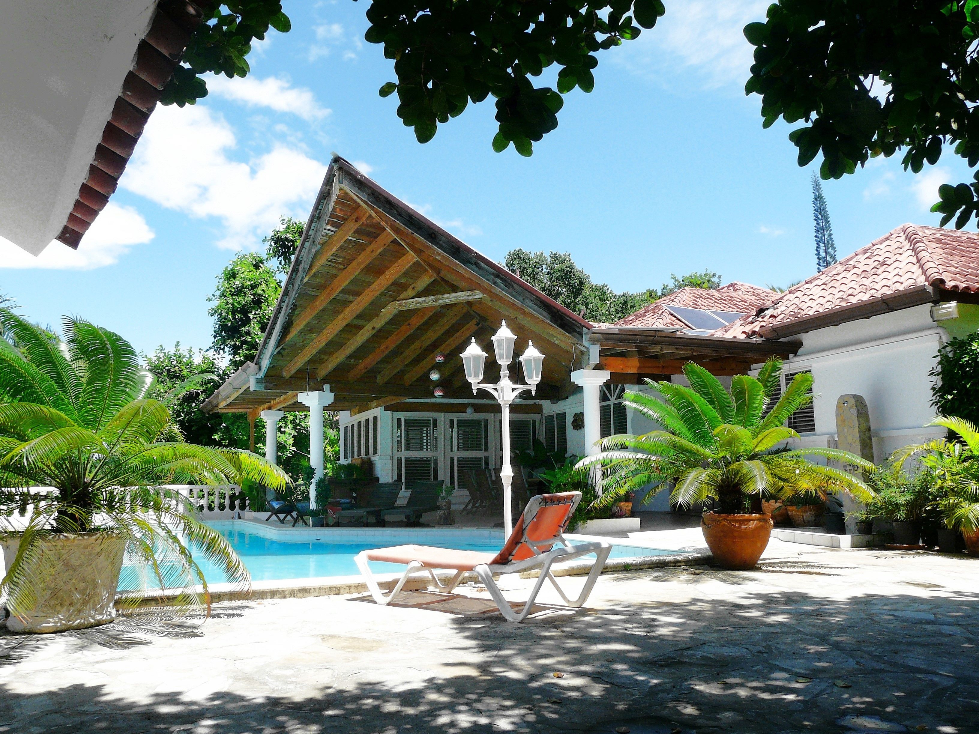 A covered pool area with a lounge chair, potted plants, and a building with a tiled roof.