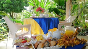 Een tafel buiten gedekt met een blauwe tafelkleed, witte stoelen en diverse etenswaren, waaronder brood, gebak en een fruitschaal.