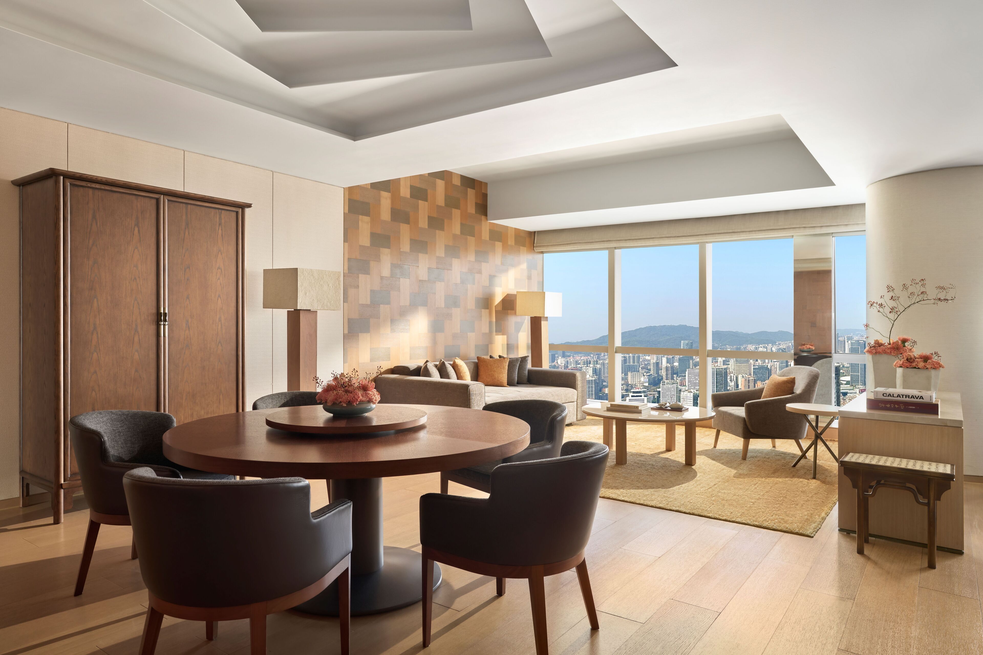 Een moderne woonkamer met een ronde eettafel, comfortabele zithoek, een groot raam met uitzicht op de stad en een houten kast.