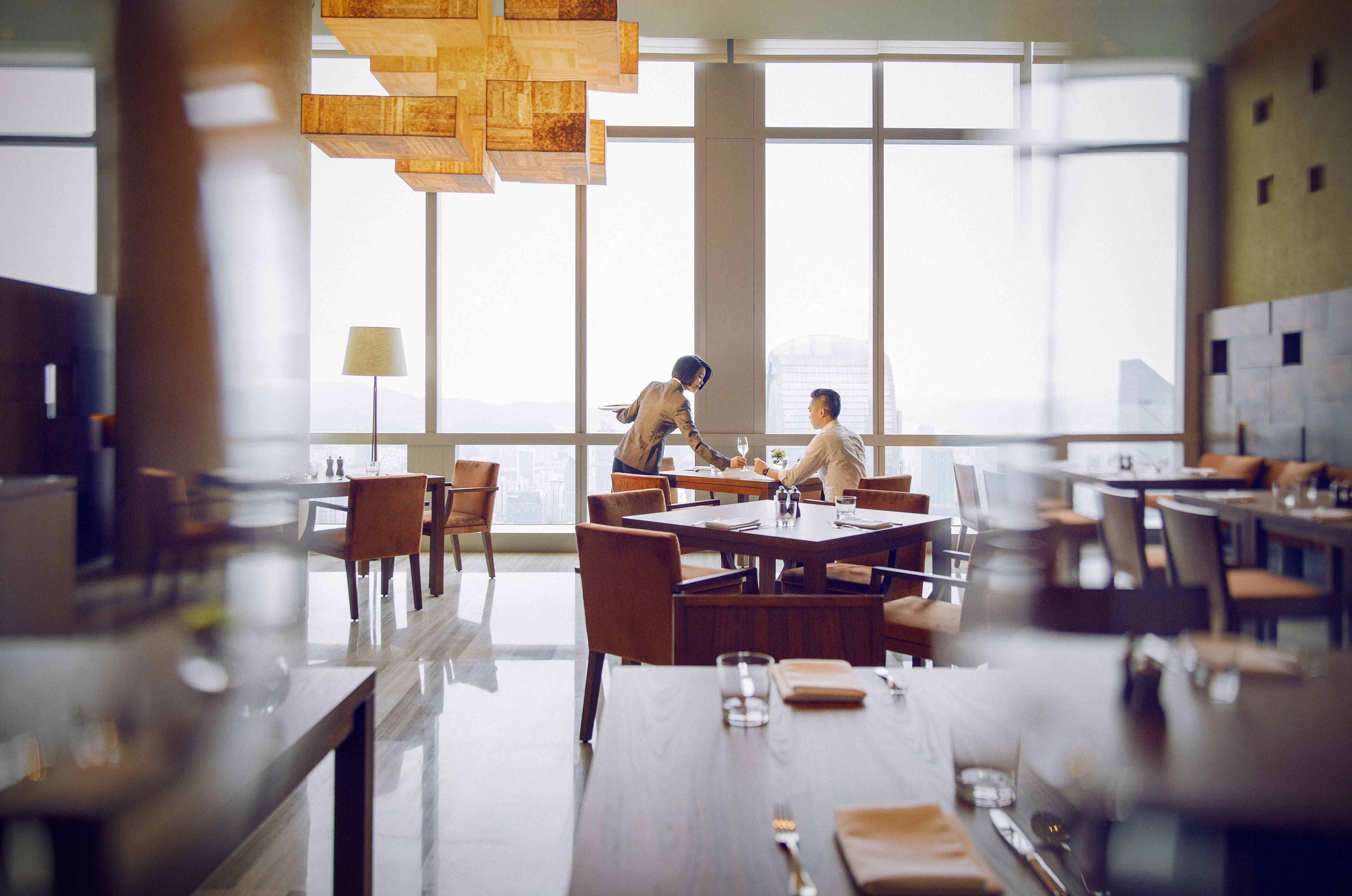 A modern dining area with large windows, wooden furniture, and a high ceiling with hanging lights.