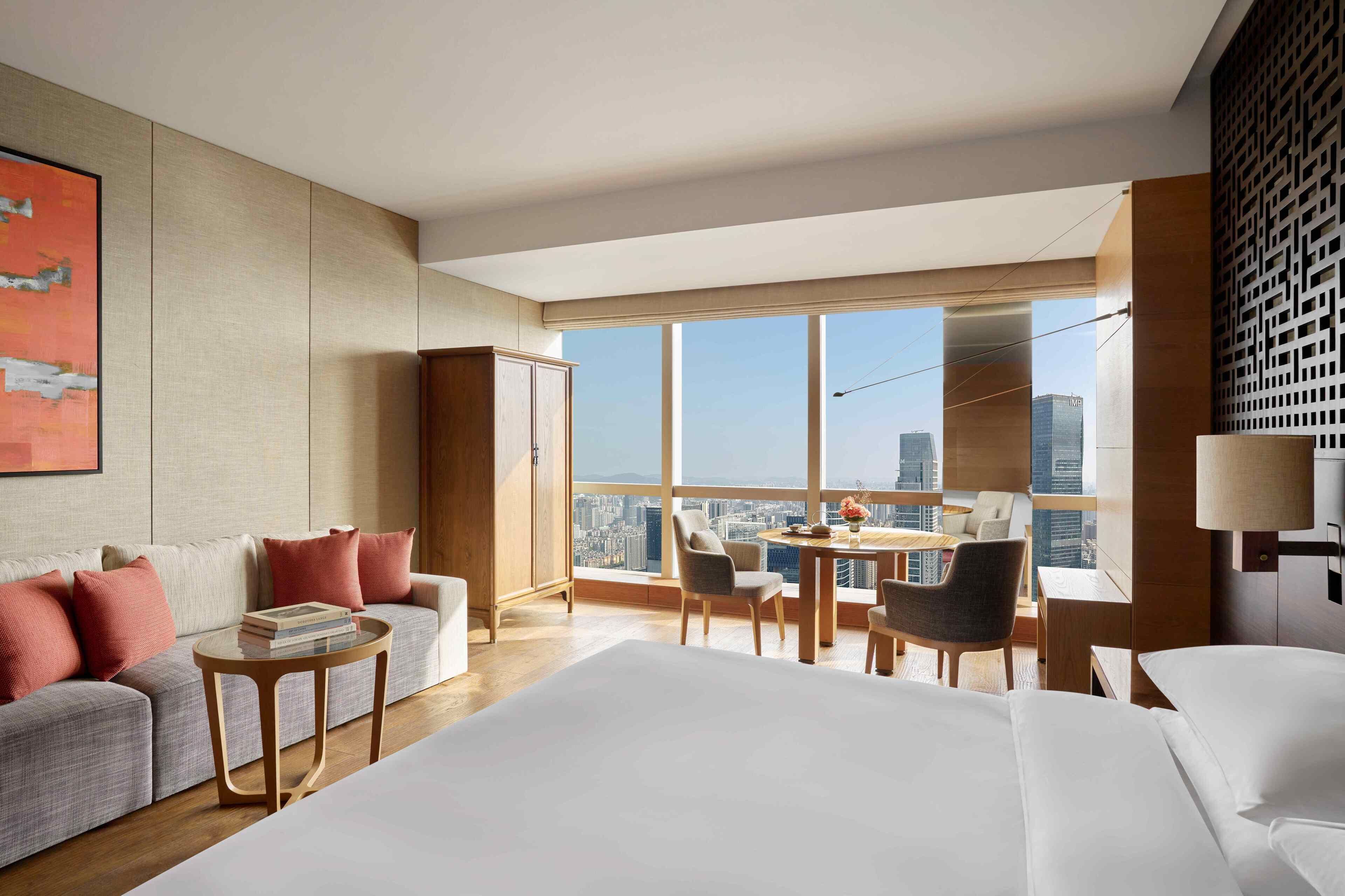 A modern hotel room with a large bed, a sofa, a dining table with chairs, and a city view through large windows.