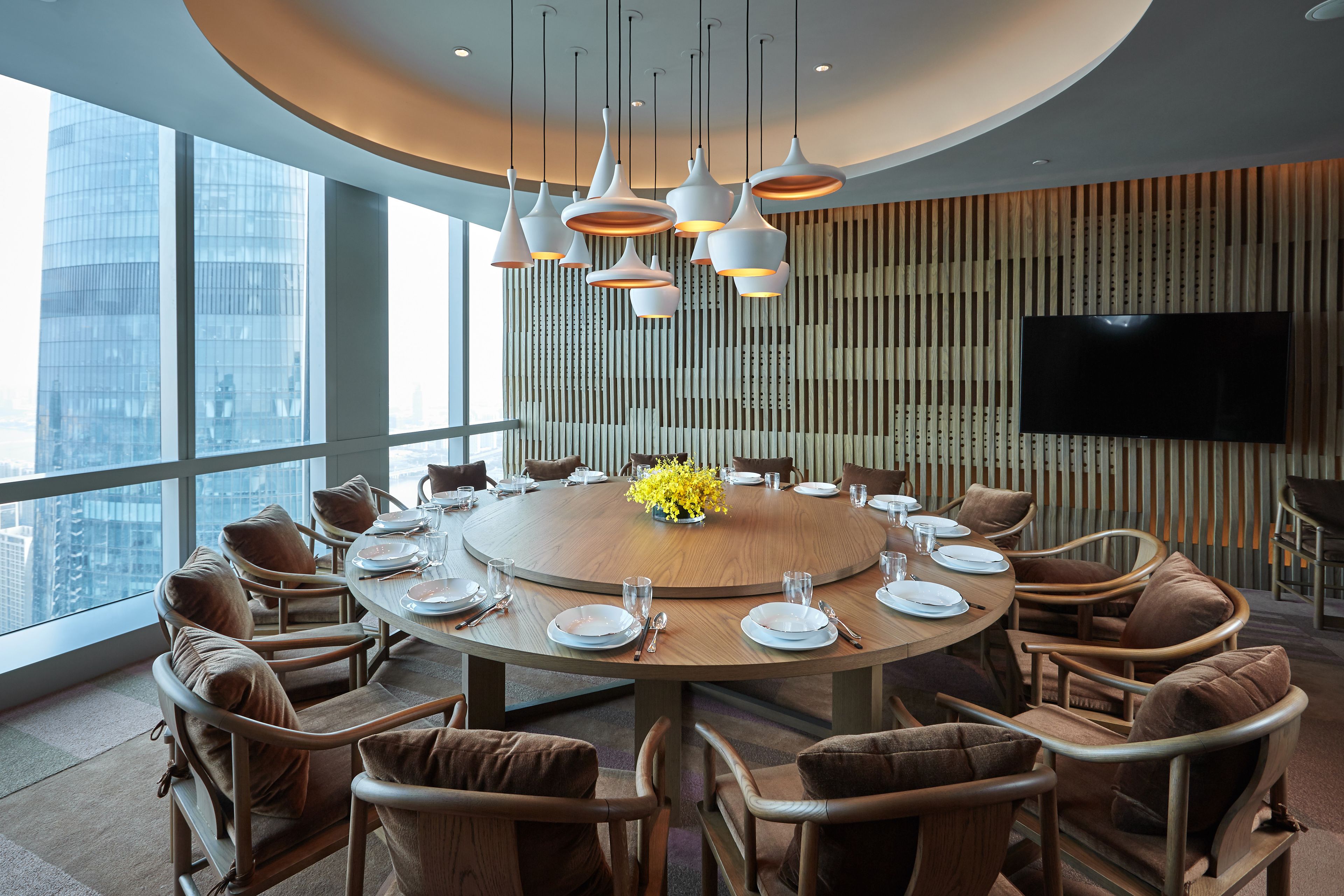 Uma sala de conferência moderna com uma mesa redonda, cadeiras estofadas em tons de castanho, um arranjo floral central e uma televisão grande na parede.