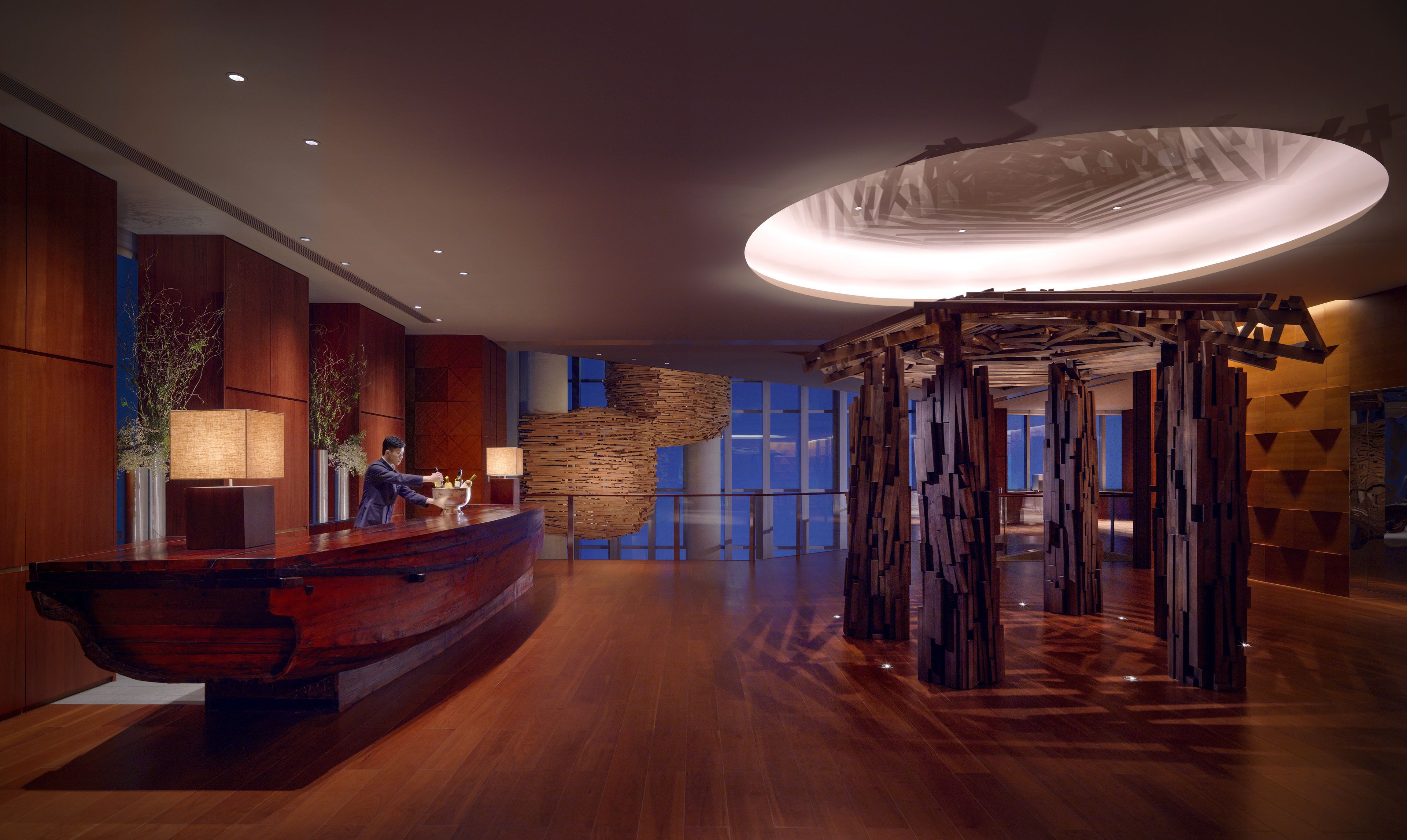 A modern lobby with a wooden reception desk, a large abstract sculpture, and a view of the sky through large windows.