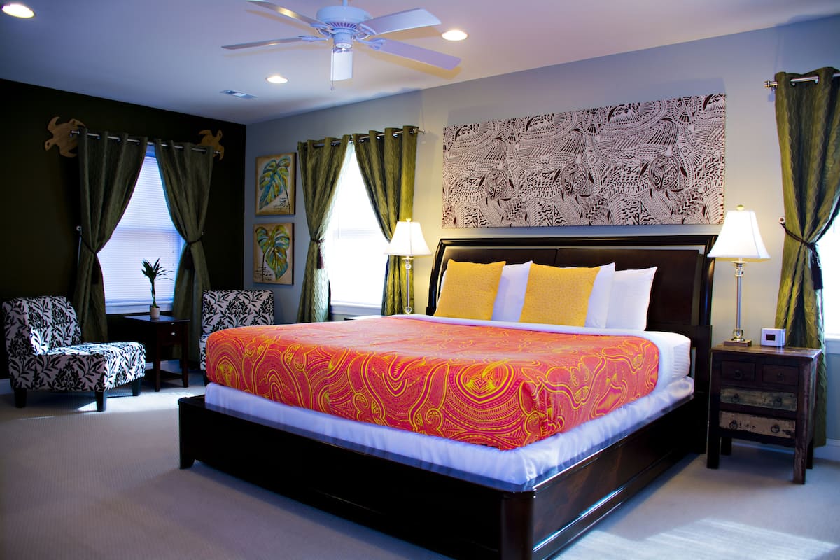 A bedroom with a large bed, a ceiling fan, a nightstand, a chair, and a window with curtains.