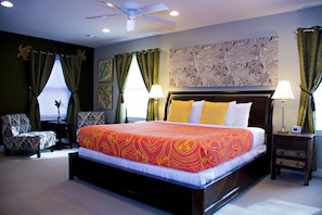 Quarto com uma cama grande, um ventilador de teto, mesa de cabeceira, uma cadeira e uma janela com cortinas.