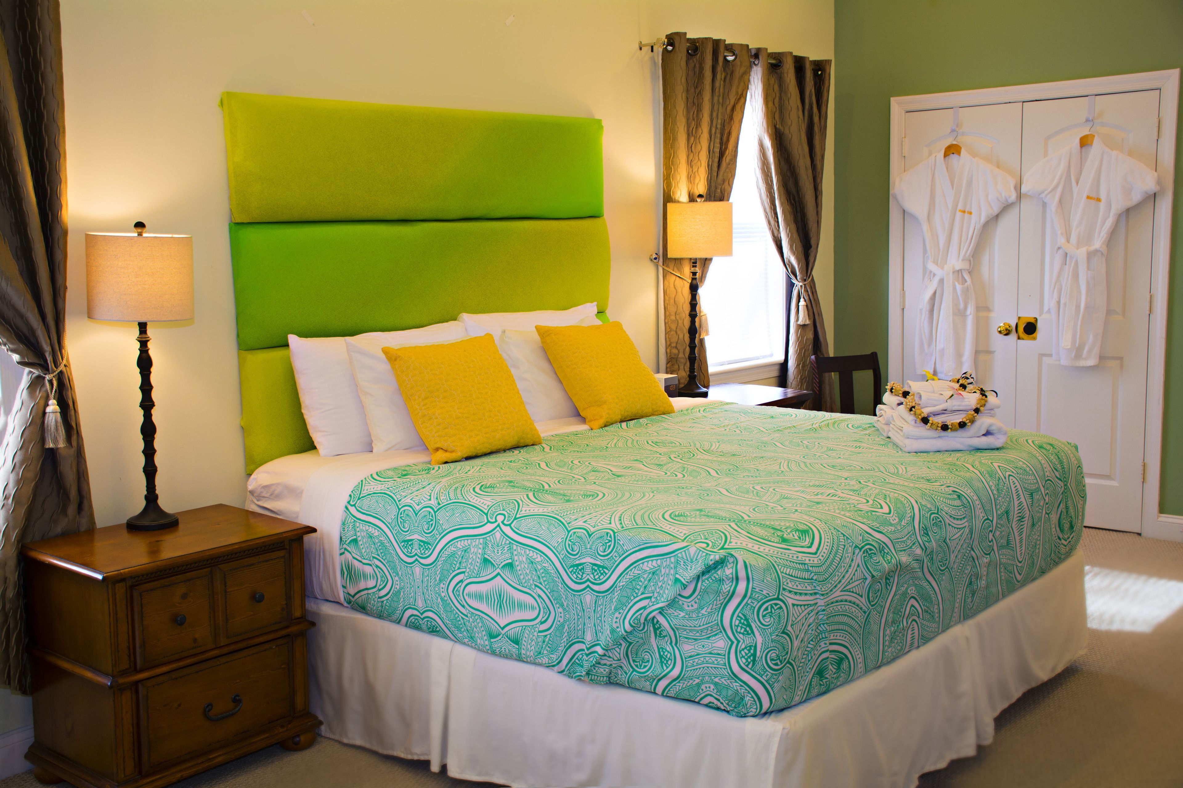 Une chambre d’hôtel avec une tête de lit verte, un lit avec une literie blanche et jaune, une table de chevet avec une lampe, et une porte sur laquelle sont accrochées deux robes blanches.