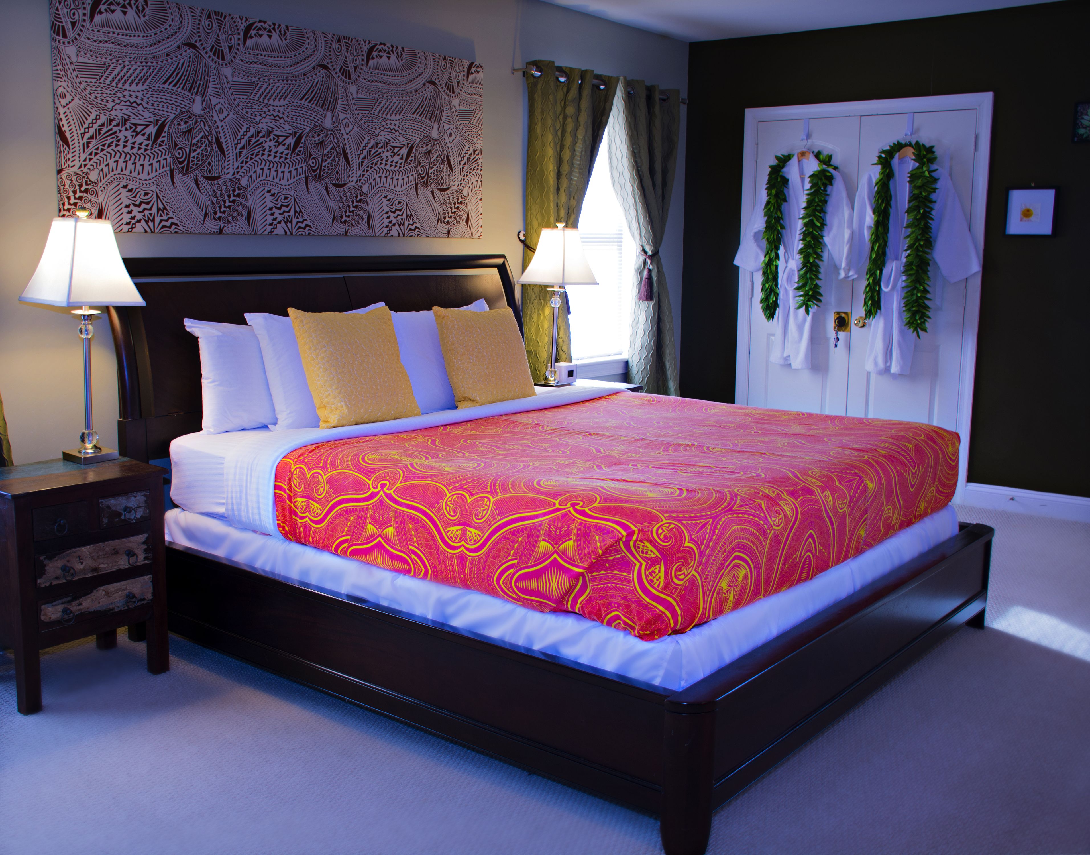 A large bed with a red and yellow patterned bedspread, two bedside lamps, a dark wooden headboard, and a wall tapestry.
