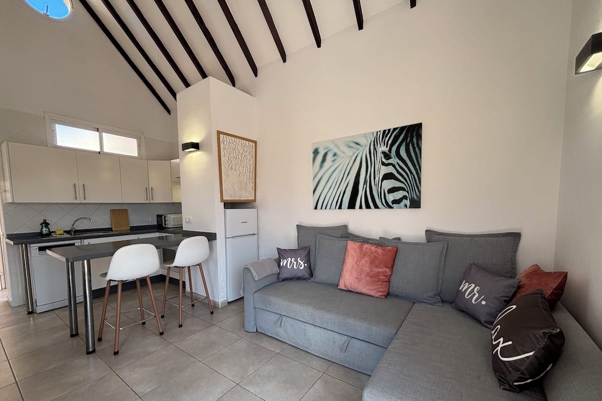A modern living room with a grey sofa, a kitchen area with white cabinets, and a dining space with two white chairs.