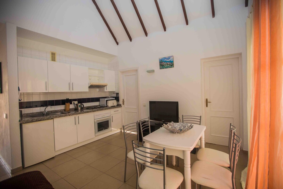 A compact kitchen with white cabinets, a dining table with chairs, a TV, and a small window with a view.