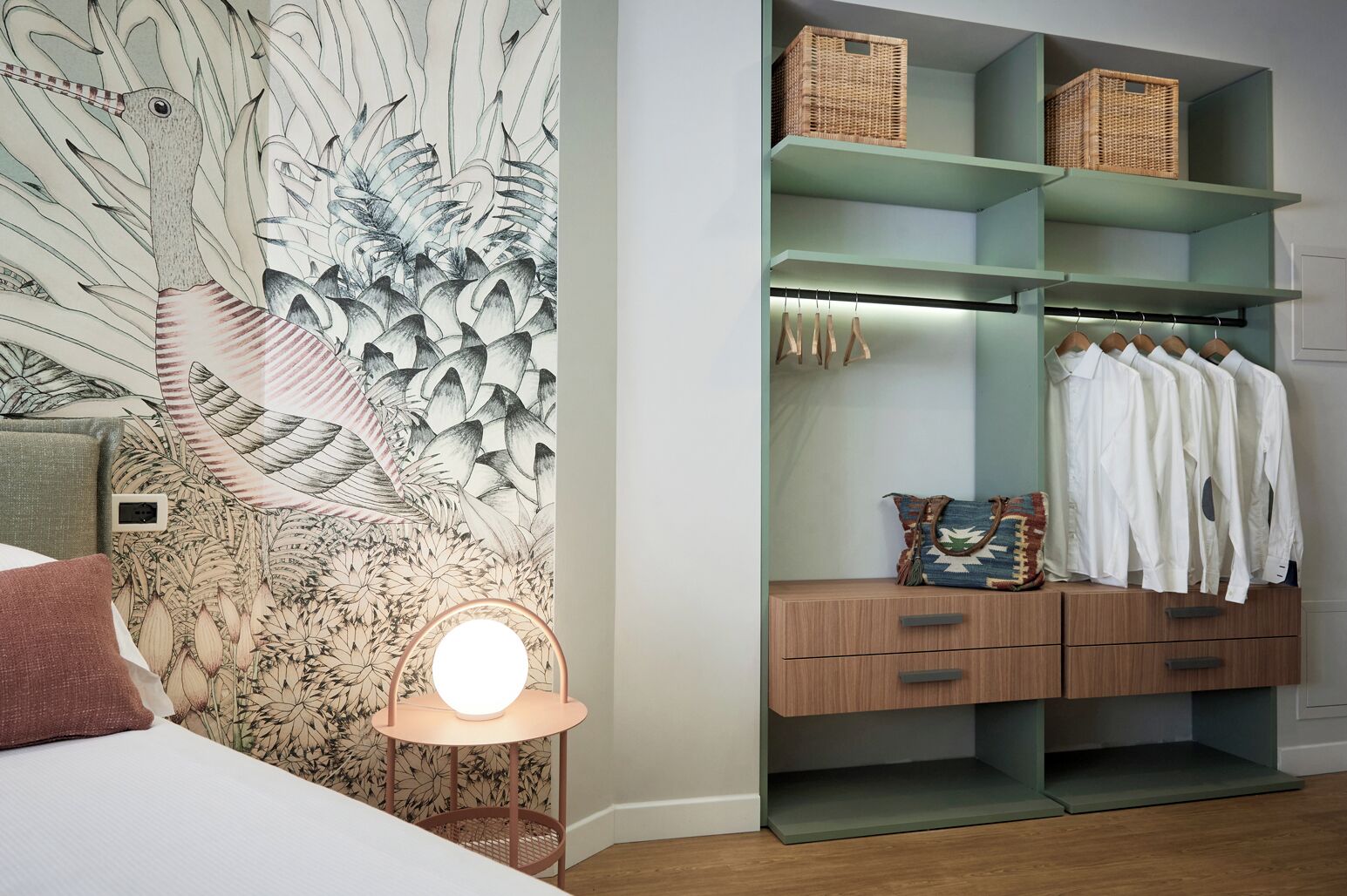 A bedroom with a large floral wallpaper, a bedside lamp, a wooden dresser, and a closet with hanging clothes and baskets.