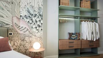 A bedroom with a large floral wallpaper, a bedside lamp, a wooden dresser, and a closet with hanging clothes and baskets.