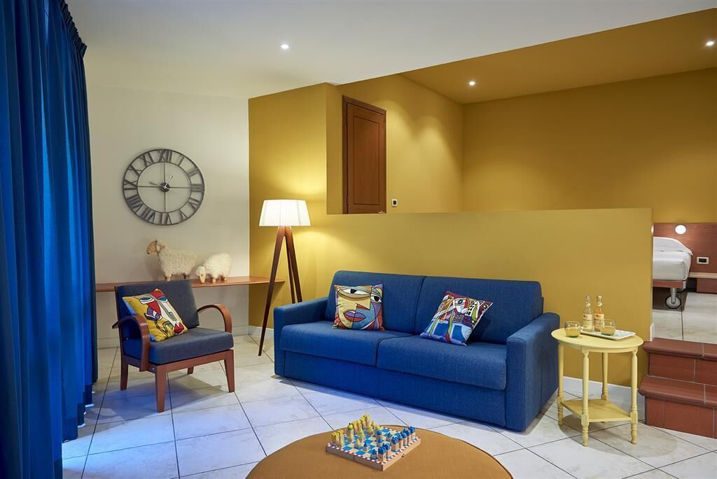 Un salon avec un canapé bleu, une chaise en bois, une table d’échecs, une lampe, une horloge et un petit chien.