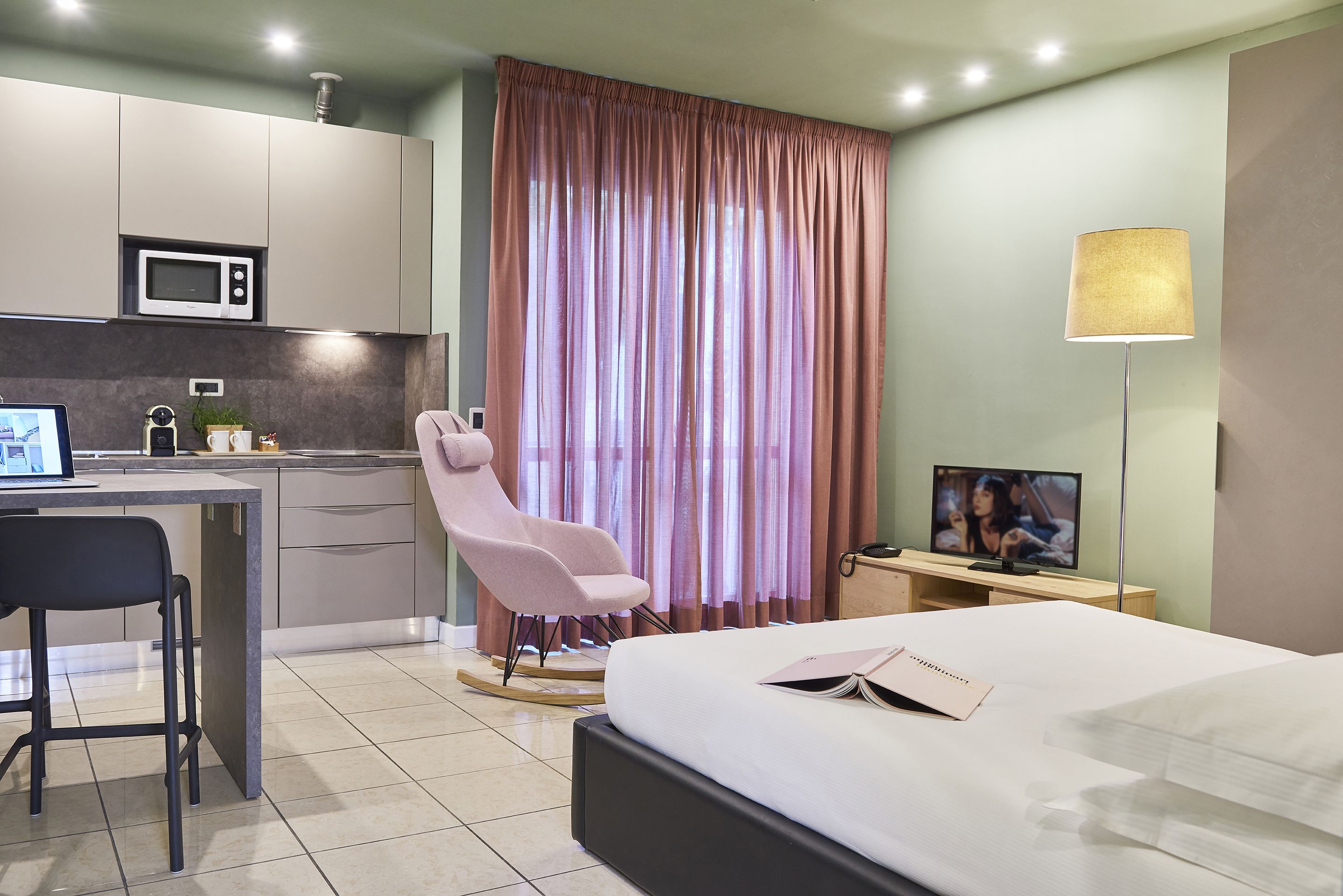 A modern hotel room with a bed, a desk with a computer, a chair, a TV, and a large window with curtains.