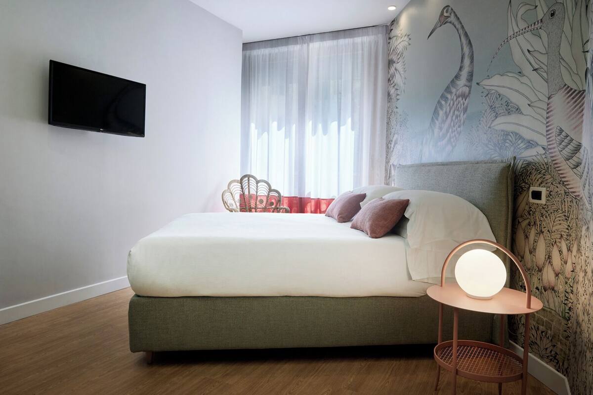 A modern bedroom with a bed, a wall-mounted TV, a bedside table with a lamp, and a decorative wall mural.