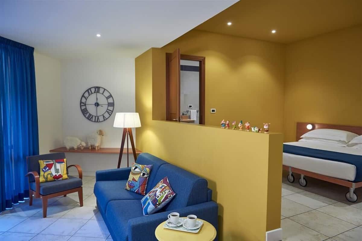 A hotel room with a blue sofa, a small round table with a tray of cups, a bed with a wooden headboard, a wall clock, and a mirror.
