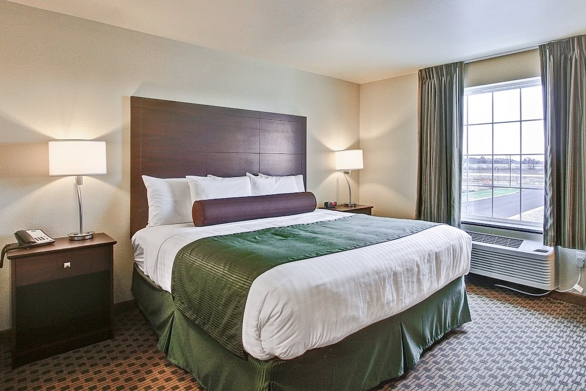 A hotel room with a large bed, two bedside lamps, a telephone, a nightstand, a window with curtains, and a radiator.