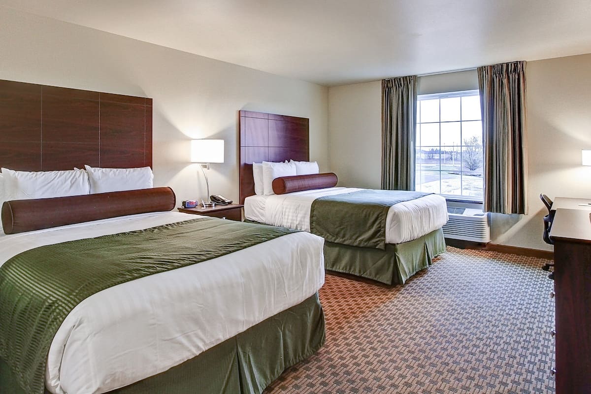 A hotel room with two beds, a desk, a chair, a lamp, and a window with curtains.