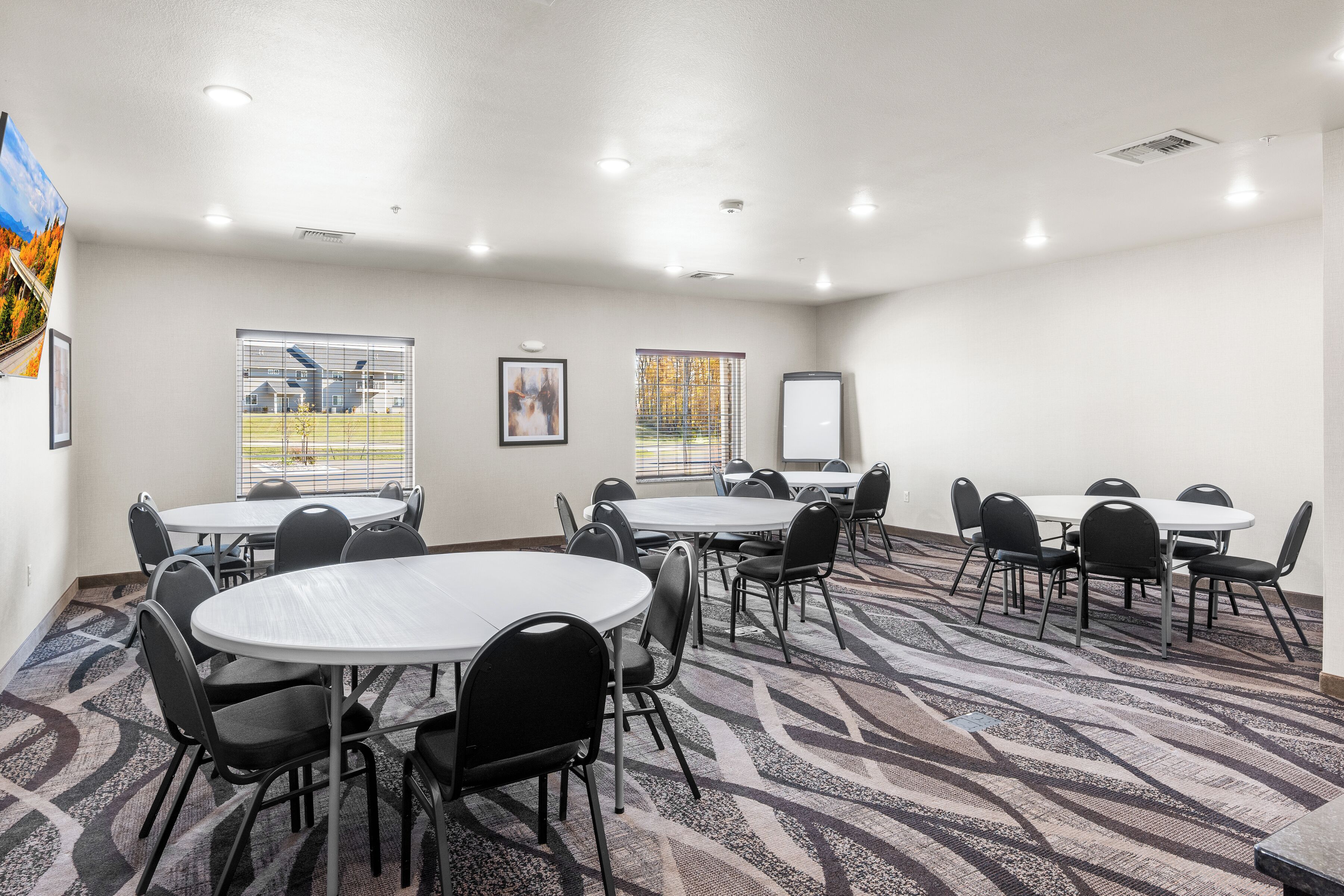 Un salón de conferencias con mesas redondas, sillas, una pantalla de proyector y ventanas con persianas.