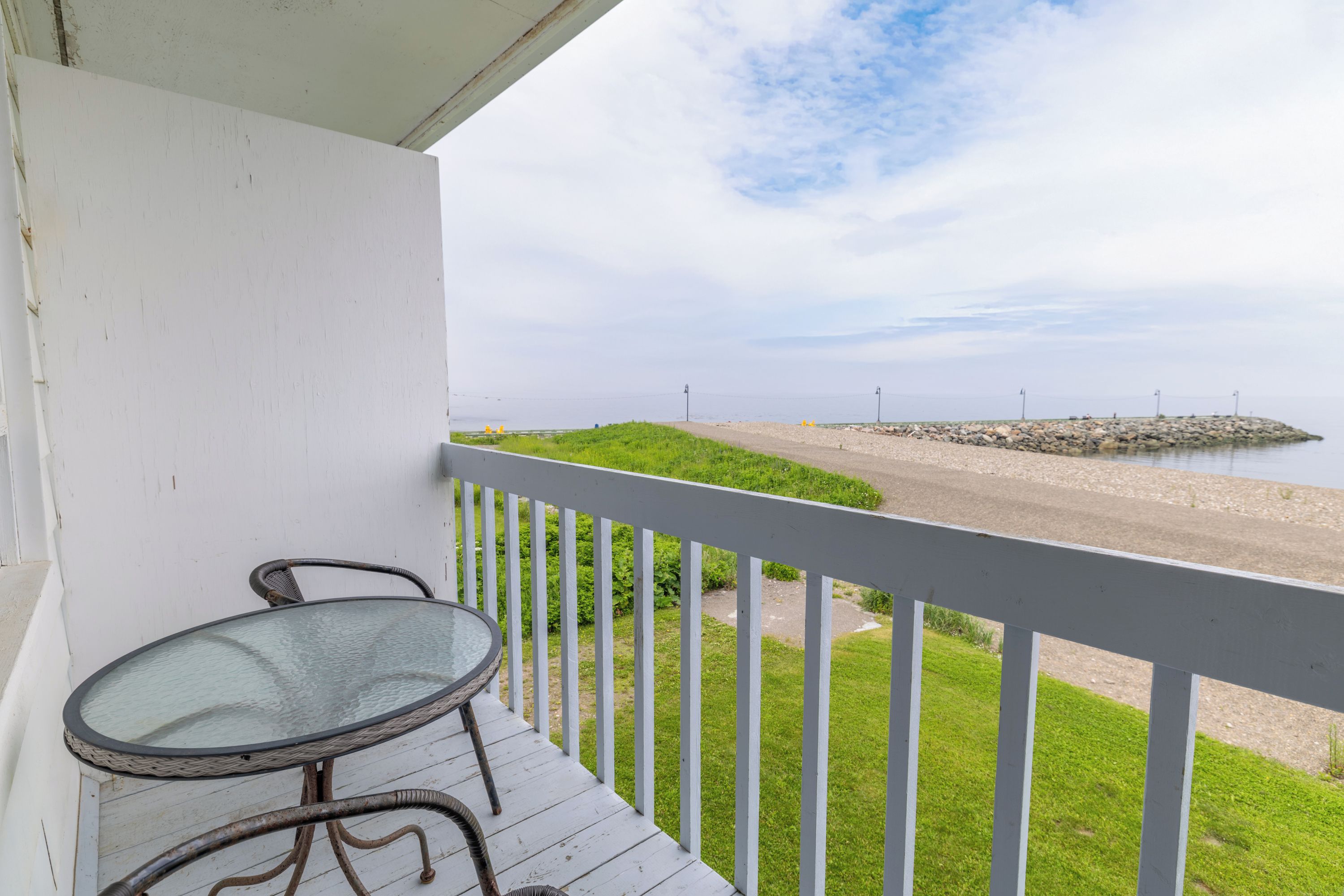Een balkon met een glazen tafel en stoelen, met uitzicht op een strand met een rotsachtige zeedijk.