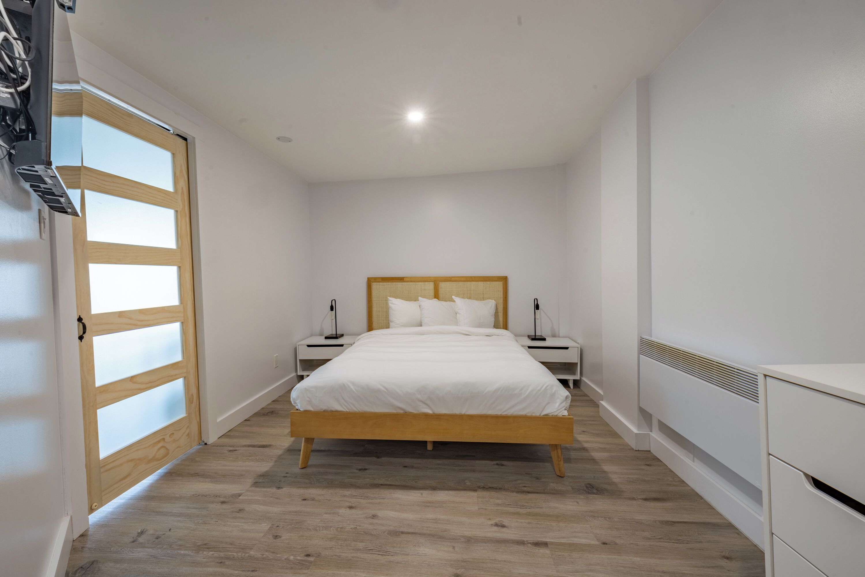 A bedroom with a wooden bed, white walls, and a wooden floor. There are two bedside tables with lamps and a white wardrobe.