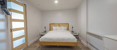 A bedroom with a wooden bed, white walls, and a wooden floor. There are two bedside tables with lamps and a white wardrobe.