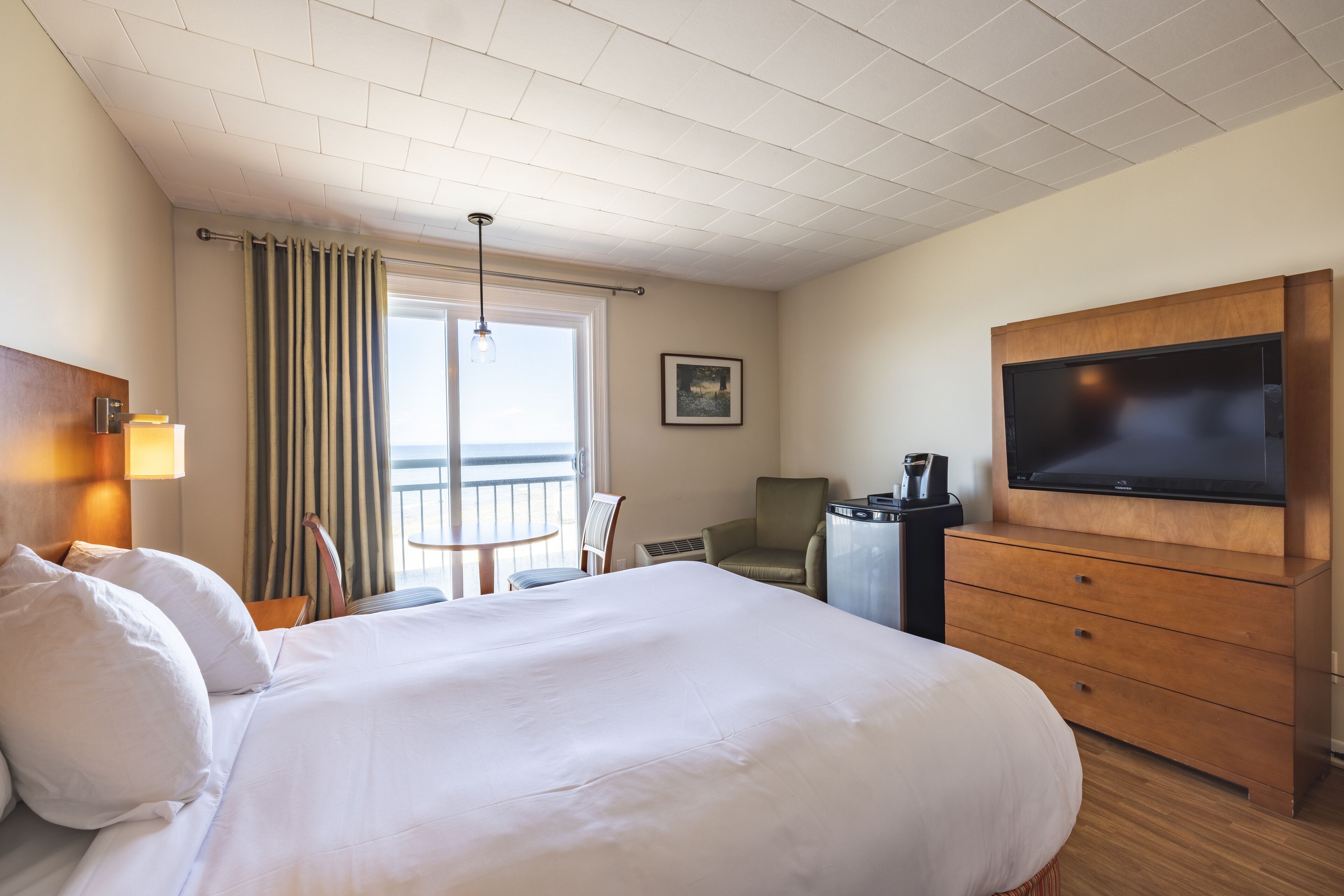 A hotel room with a large bed, a TV, a desk with a coffee maker, a chair, and a view of the outdoors.