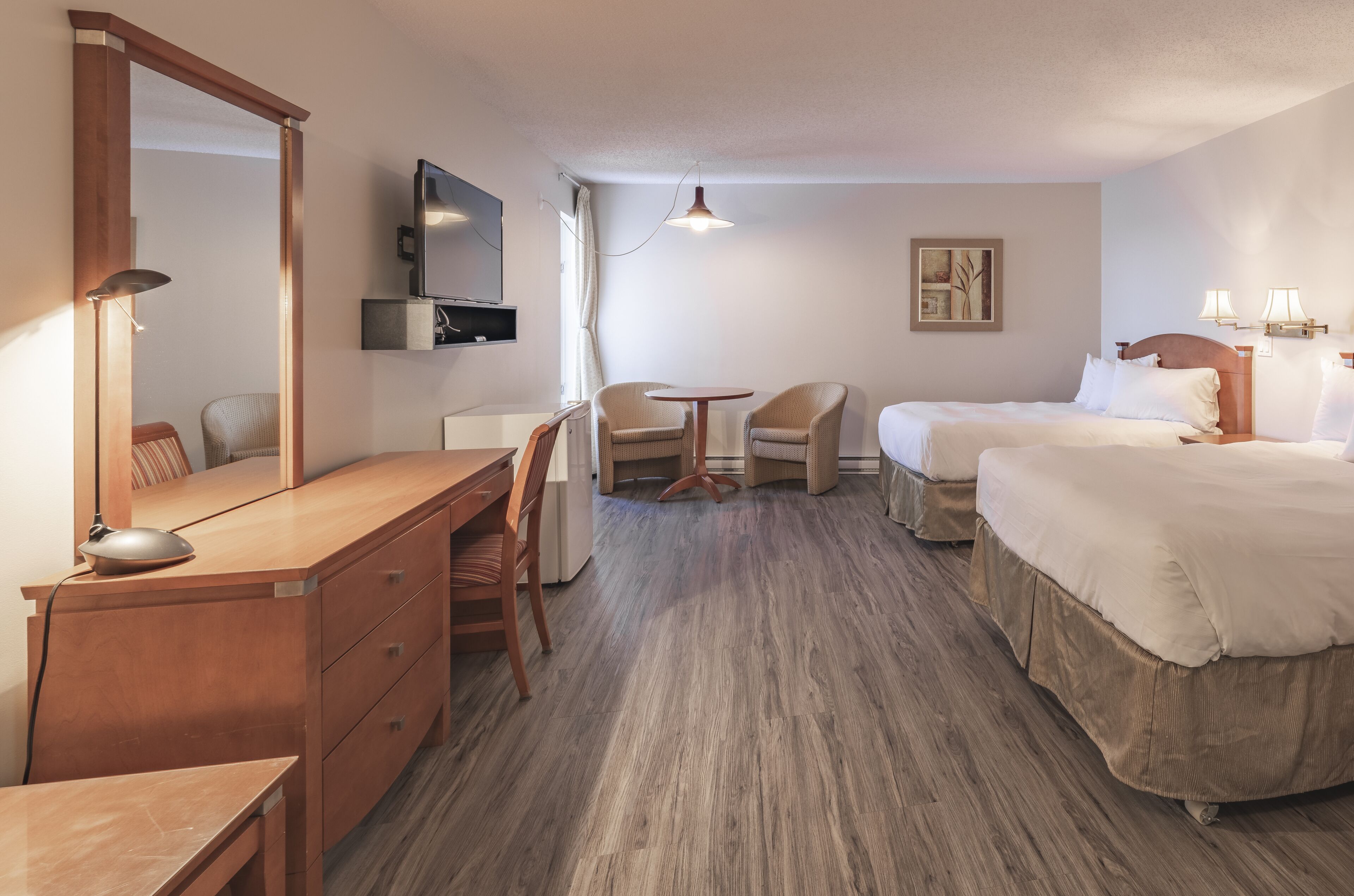 A hotel room with a wooden desk, two single beds, a round table with chairs, a TV, and a ceiling light.