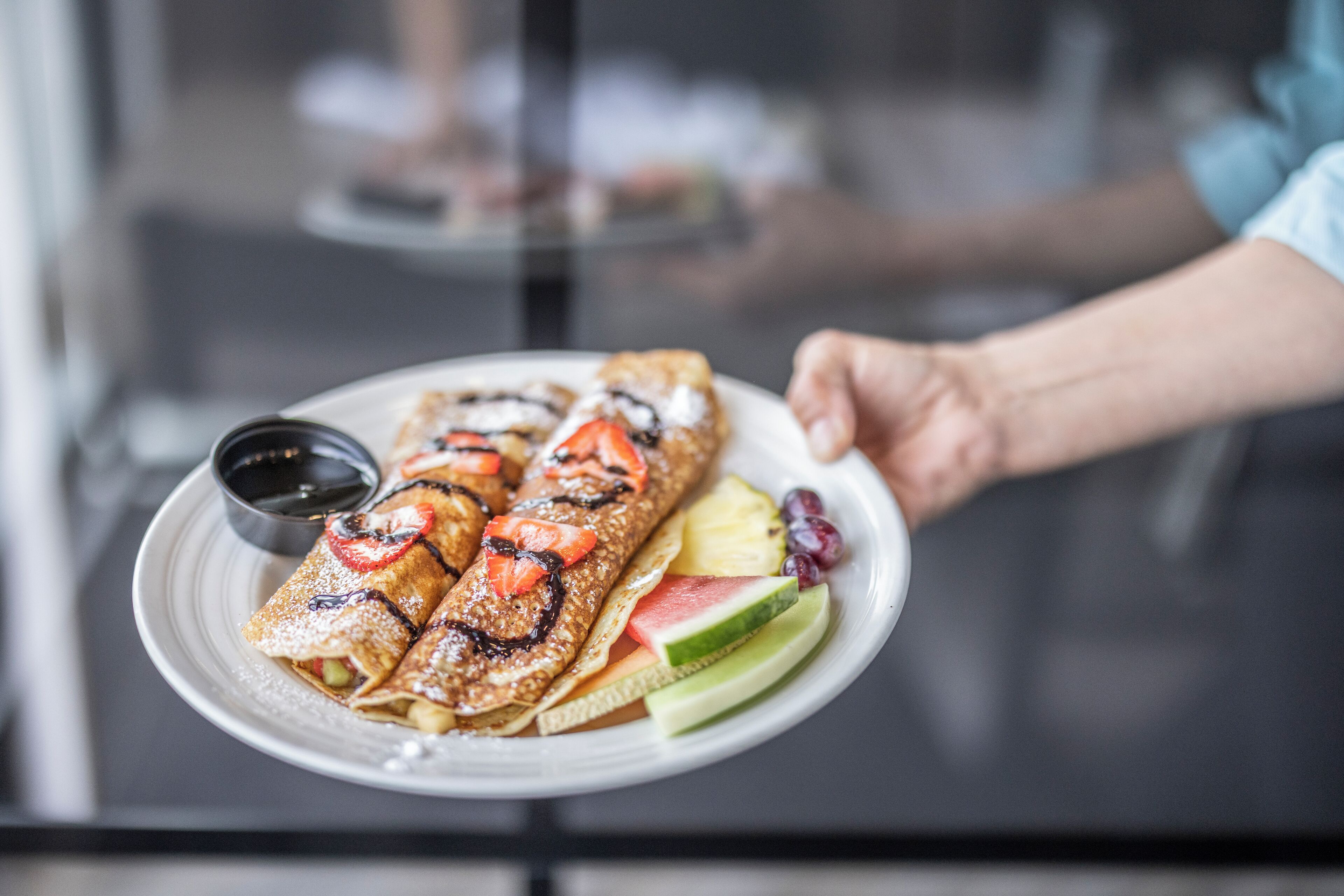 A plate with crepes, fruit, and a small cup of syrup.