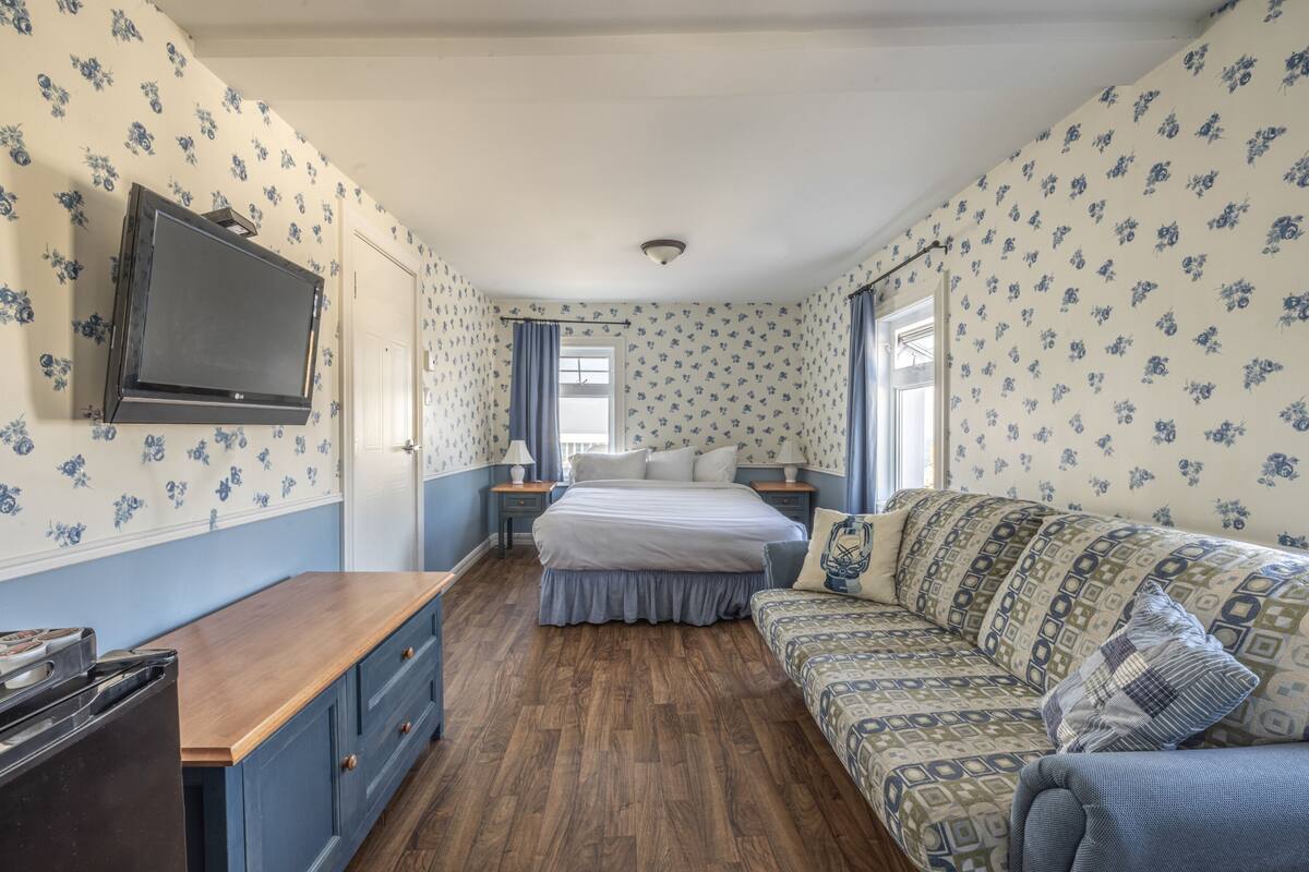 Ein Schlafzimmer mit Holzboden, einem Bett mit weißer Bettwäsche, einem blau gemusterten Sofa, einem an der Wand montierten Fernseher und einem Fenster mit Vorhängen.