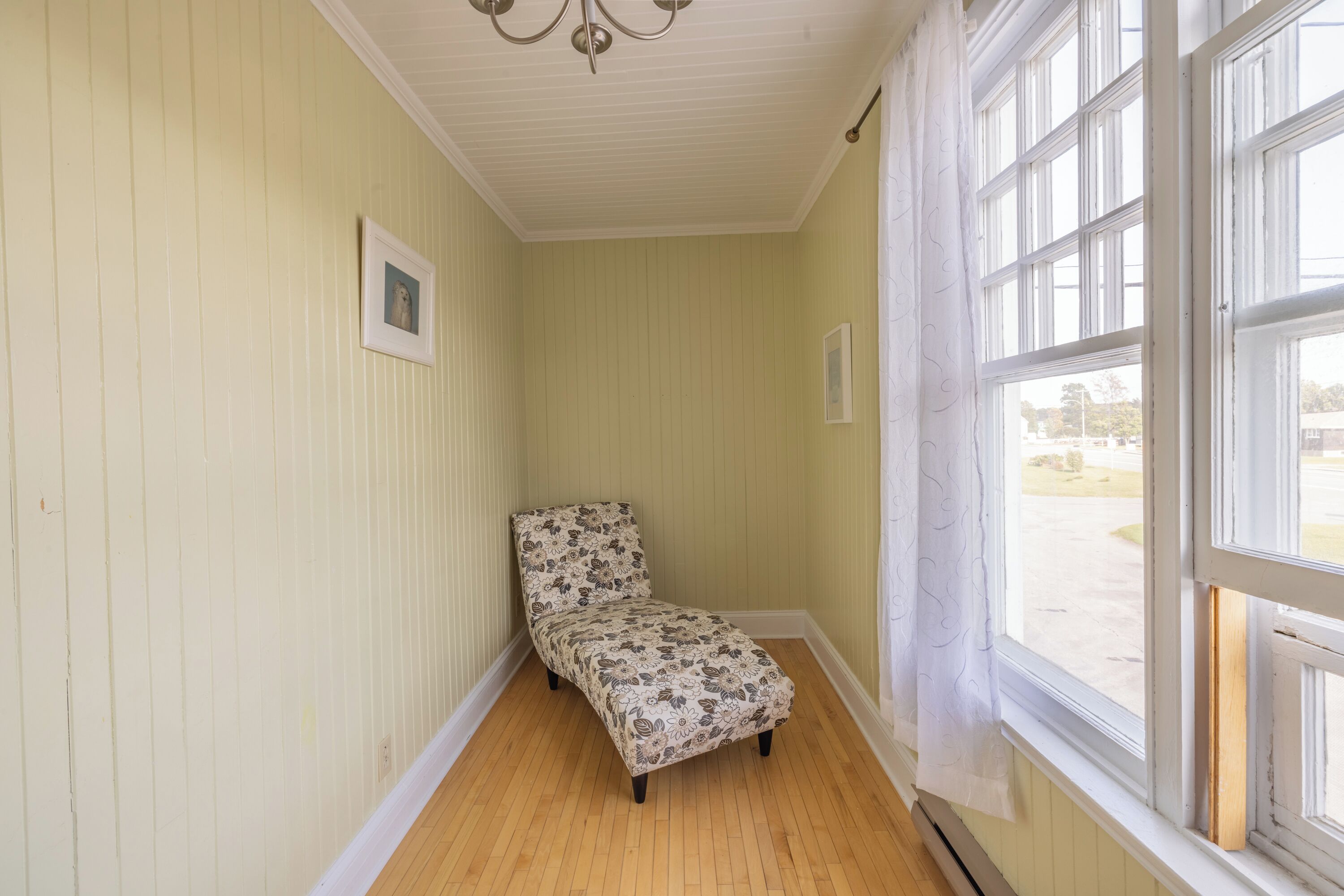 A narrow hallway with a single chair, wooden flooring, and a large window with a view of the outdoors.