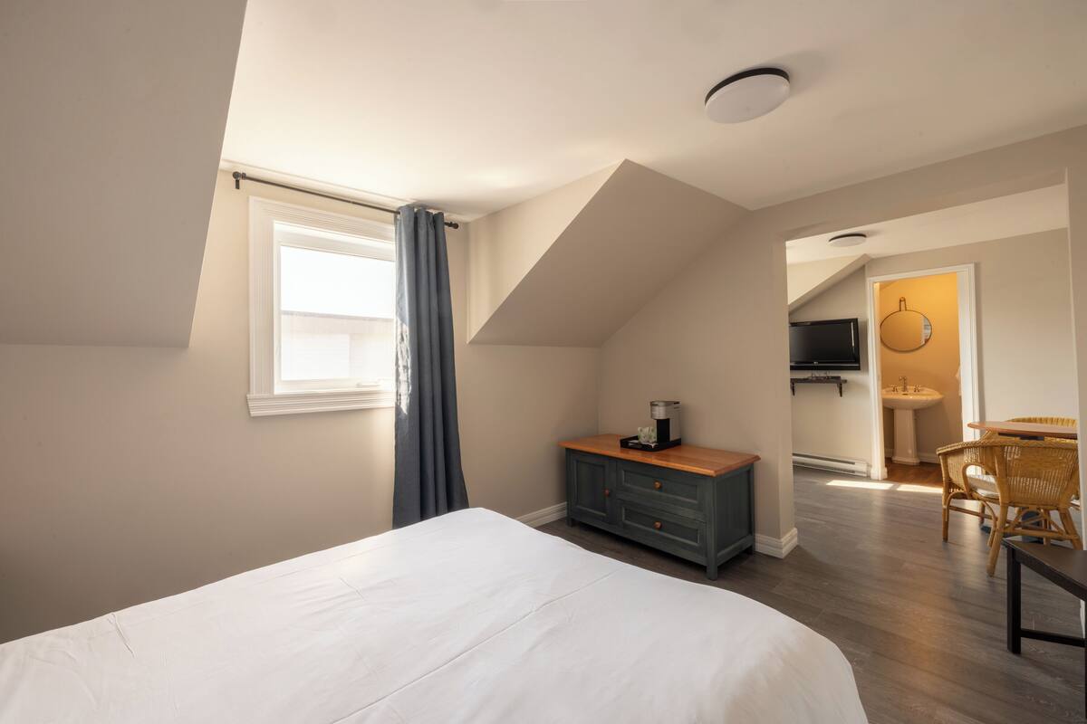Ein Schlafzimmer mit einem großen Bett, einem Schminktisch, einem Fenster mit Vorhängen und Blick in einen Küchenbereich.
