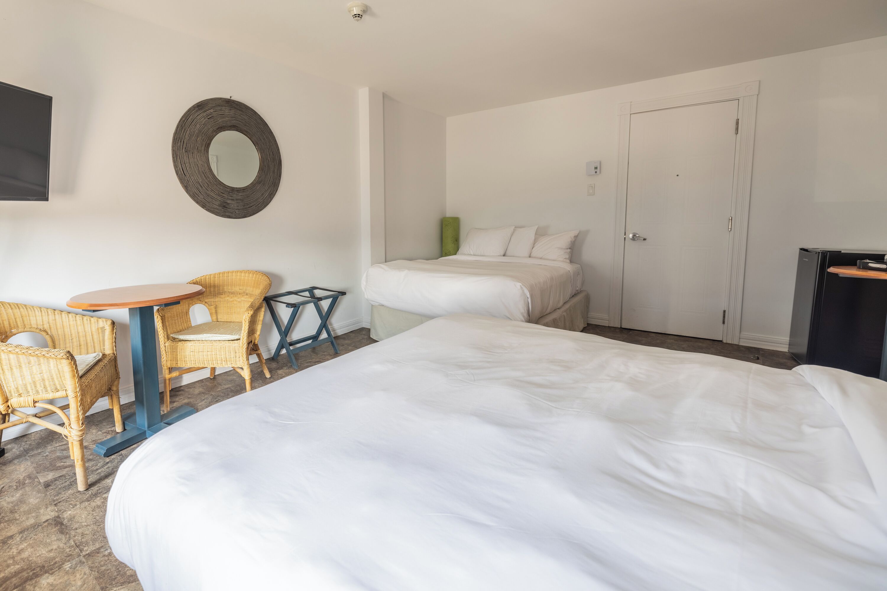 A hotel room with two beds, a television, a round mirror, wicker chairs, and a small table.