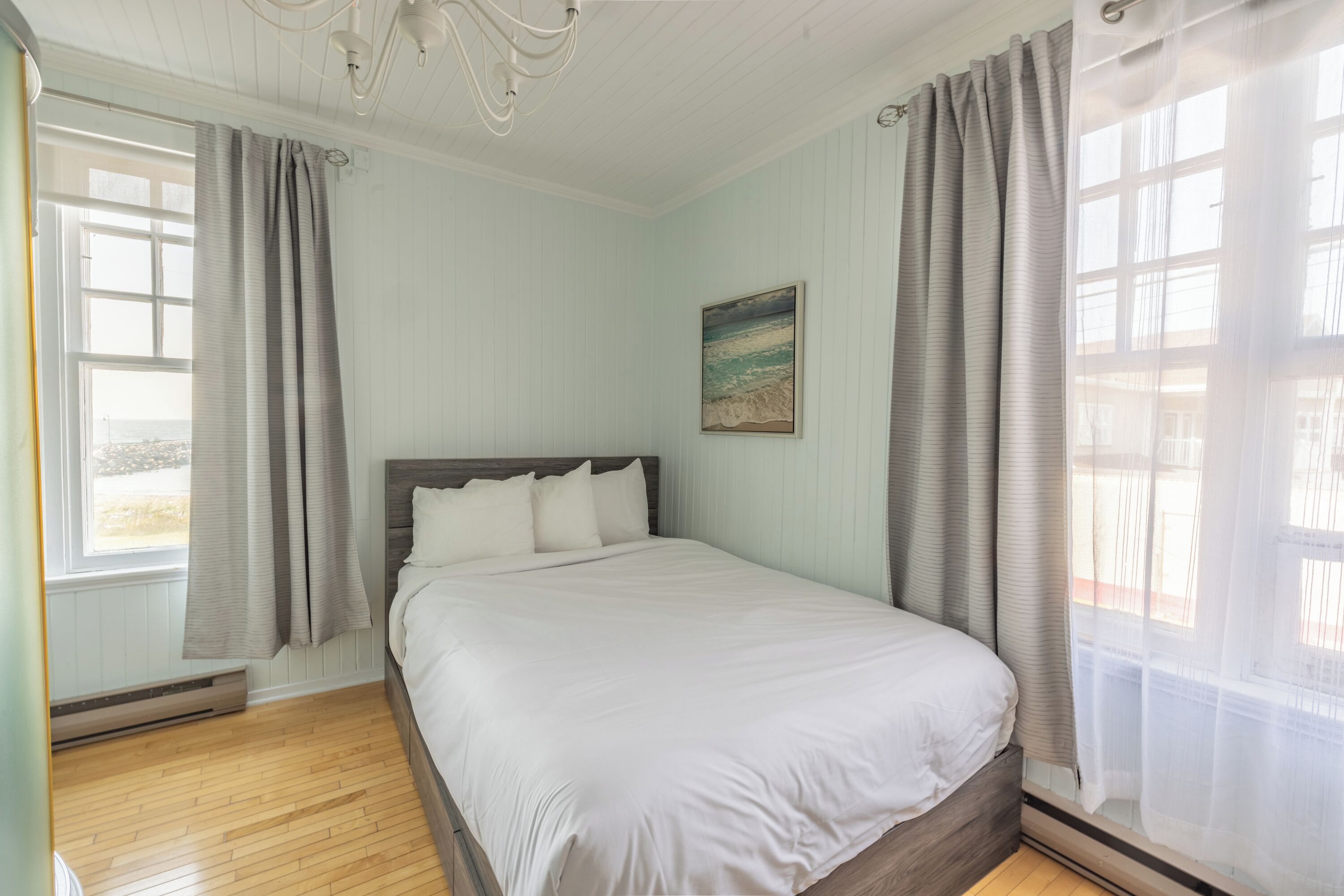 A bedroom with a wooden floor, a bed with white bedding, a painting on the wall, and a window with curtains.