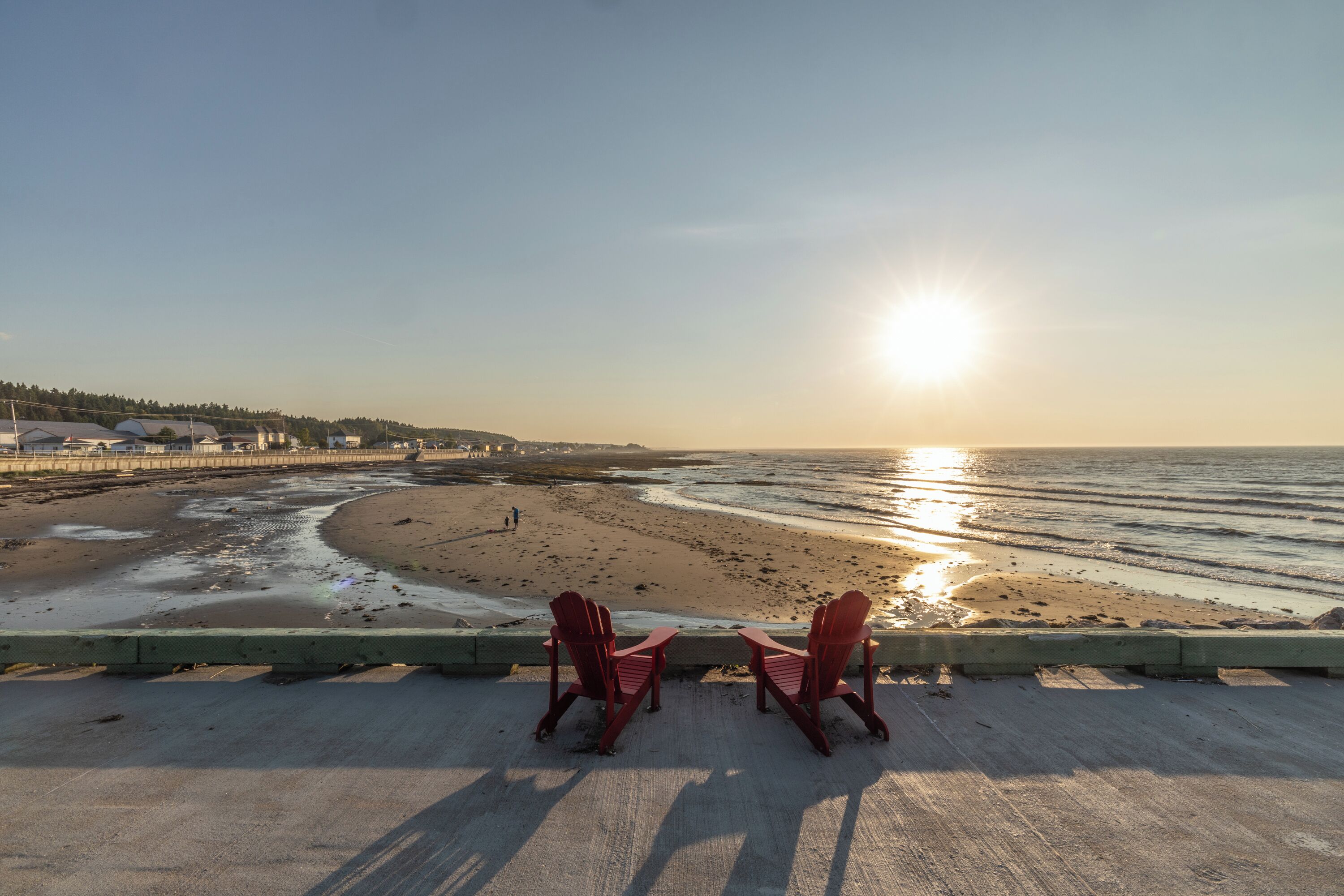Twee rode Adirondack-stoelen gericht op een strand, met een ondergaande zon op de achtergrond.