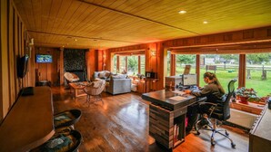 Un vaste espace de vie doté d’un plafond en bois, d’une cheminée, d’une grande fenêtre et d’un coin repas avec une table et des chaises.